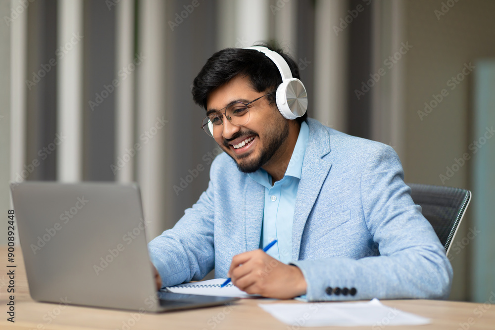 Aufkleber A smiling Indian man wearing headphones sits at a desk and works on a laptop computer. He is taking notes with a pen on a notepad and smiling. There are papers and a pen on the desk.