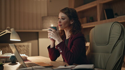 Pensive manager drinking tea sitting dark office desk with laptop close up. 