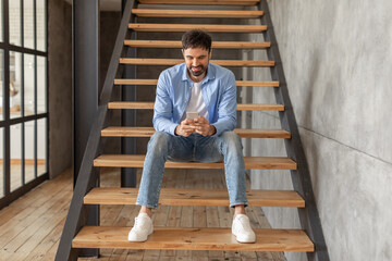 A man sits on a set of wooden stairs in a modern home. He wears a blue shirt, white t-shirt, blue...