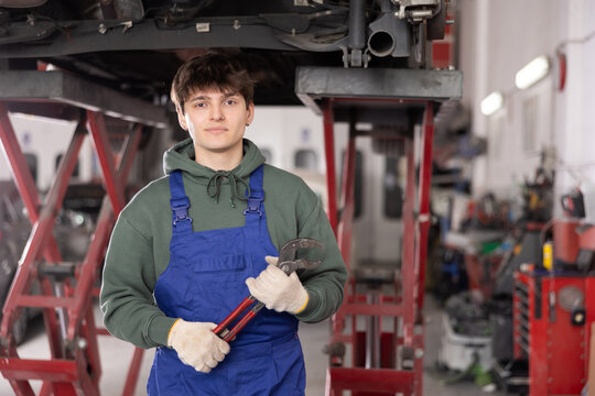 Positive young mechanic in blue overall, holding adjustable wrench, standing under car raised on hydraulic lift in professional well-equipped vehicle repair shop..