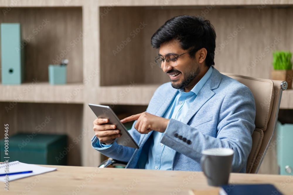 Leinwandbilder a smiling indian businessman sits at a desk in his office, looking at a tablet and using his finger 