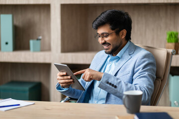 A smiling Indian businessman sits at a desk in his office, looking at a tablet and using his finger to navigate the screen. There is a cup of coffee on the desk and a notebook with a pen beside it.