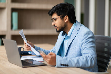 Fototapeta premium A young Indian man in a blue blazer sits at a desk, looking intently at a document. He has a laptop open in front of him, and his other hand is holding a smartphone.