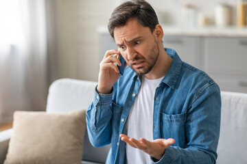 A man with a concerned expression sits on a sofa in a living room, talking on a mobile phone. He appears frustrated, with one hand raised in the air, as he speaks.