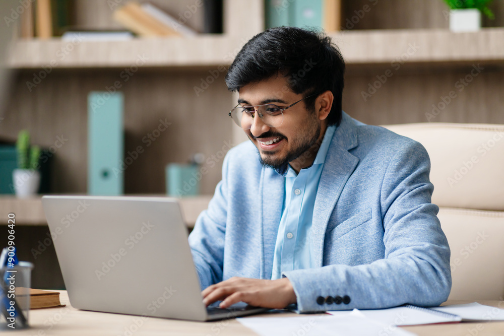Aufkleber indian man wearing a light blue jacket sits at a desk in a modern office, smiling as he works on his