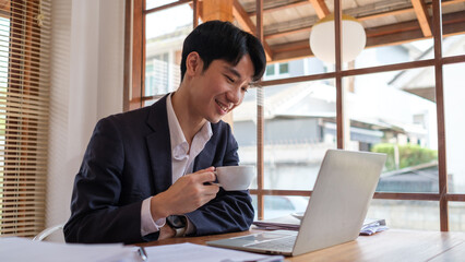 Young businessman in a suit is smiling as he enjoys a coffee break while working on his laptop in a modern office.