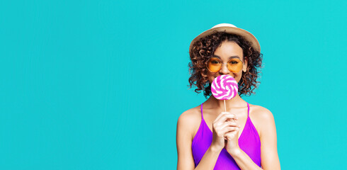 A young woman with curly hair, wearing a purple swimsuit, sunglasses, and a straw hat, smiles while holding a pink and white lollipop in front of her face. She stands against turquoise background.