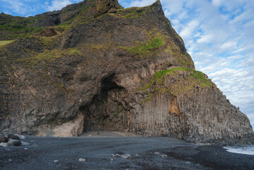 Reynisfjara Beach in Iceland features striking basalt columns and cave formations. The dark volcanic sand contrasts with moss-covered cliffs under a partly cloudy sky.
