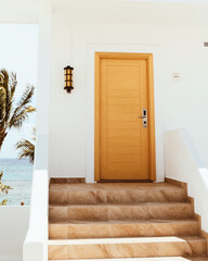 A minimalist and inviting entrance to a seaside villa in Cancun, Mexico. The light wooden door and white walls are complemented by a small staircase and tropical palm trees