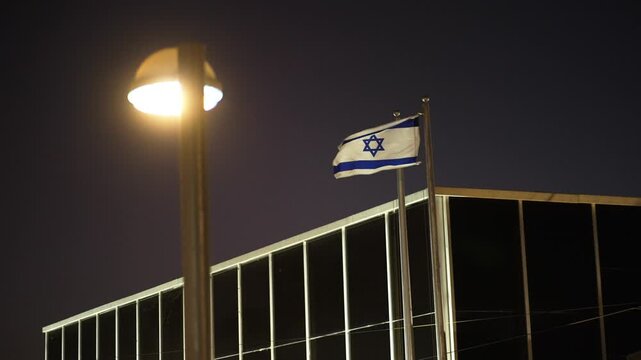 The Israeli flag flutters in the wind
Israeli flag white and blue with star Magen David fluttering in wind, Above the roof of a building at night.
A street lamp in front of the frame