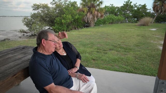 senior citizen married couple sit on a park bench near the ocean talking and enjoying life