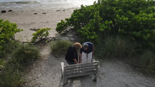 older couple sit down at the beach to rest on a bench to watch the sunset