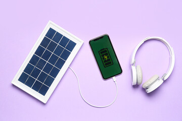 Portable solar panel charging mobile phone and headphones on lilac background