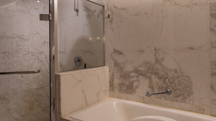 A fragment of the bathroom. The shower cabin is fenced with glass partitions. A white rectangular bathtub in the corner. A metal handrail on a beige tiled wall.