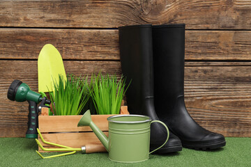 Composition with watering can, rubber boots and water sprayer on green grass against wooden background