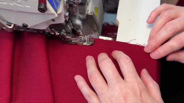 A close-up view of hands as they guide piece of red fabric through serger machine. machine as machine processes edge of the cut creating clean and professional finish fabric is folded over on itself.