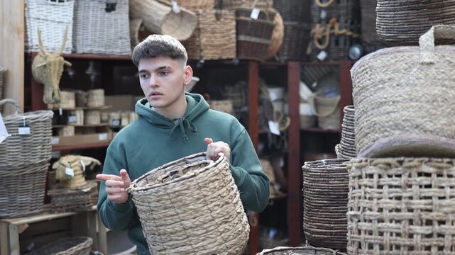 Young man buyer in store of homemade self-made goods made of osier shoot, wicker decorative items considers large and small rattan baskets for storing items. Modern design and natural materials