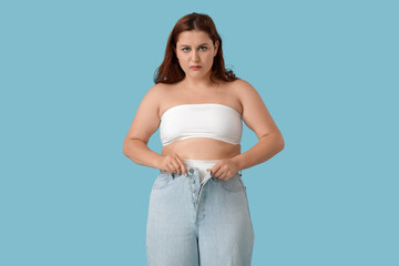 Overweight young sad woman in tight jeans on blue background. Weight gain concept
