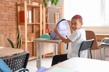 Little African-American schoolboy with backpack sitting at desk in classroom