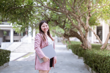 Businesswoman, standing holding a laptop, she looks happy with something, behind her are many tall trees