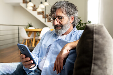 Mature man relaxing at home, sitting on the sofa sending text message with mobile phone.