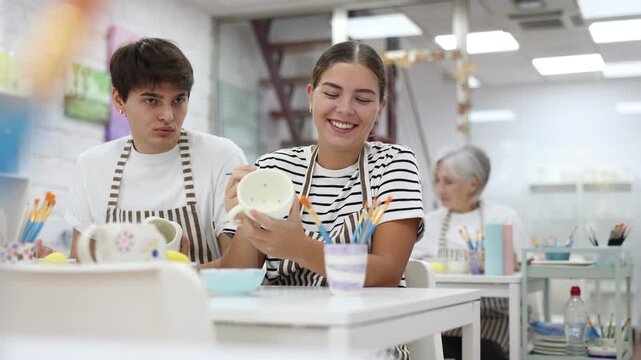 Young couple joyfully talking and working together on ceramic pottery, enjoying creative process while painting mugs in light workshop