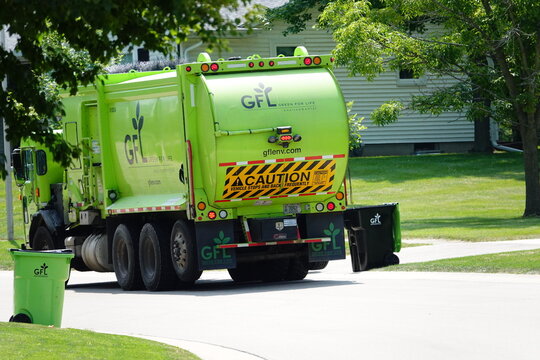 Green GFL garbage truck picking up garbage containers on the side of the residential street.