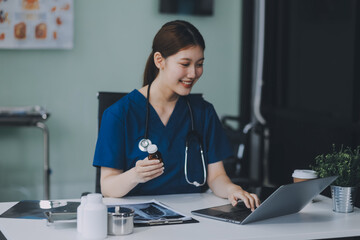 Female doctor writing in a notebook and holding a medicine bottle works at a computer while there were many medicine bottles on the table while sitting at a work desk