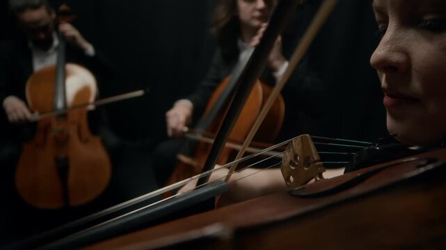Elegant string quartet sitting on chairs next to each other on dark stage with black curtains and playing violins and cellos