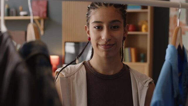 Chest up of pretty Biracial girl looking through clothes hanging on rack in living room and choosing ootd then smiling at camera
