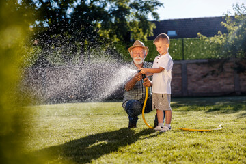 Happy child and his granddad watering lawn in backyard with gardening hose.