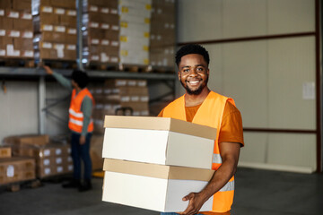 Happy diverse warehouse worker carrying boxes in hands and smiling at camera.