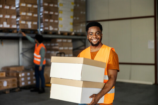 Happy diverse warehouse worker carrying boxes in hands and smiling at camera.