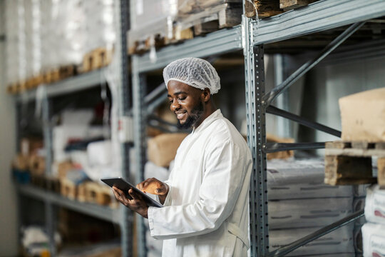 Side view of happy diverse food factory supervisor standing in warehouse and tracking goods and shipment on tablet.