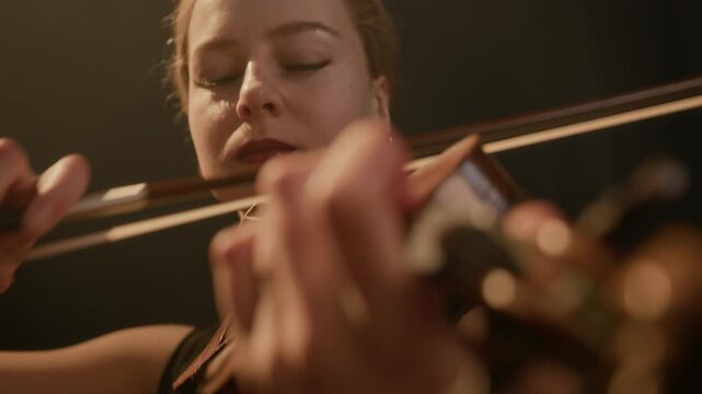 Medium closeup of graceful young Caucasian female violinist playing with expression on dark stage