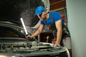 A mechanic wearing a blue shirt and overalls inspects a car engine with a flashlight. He is...