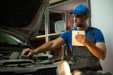 A mechanic wearing a blue shirt and grey overalls is using a tablet to diagnose a problem with a...