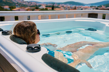 Woman wearing sunglasses and swimsuit relaxing in outdoor hot tub with bubbling water and enjoying...
