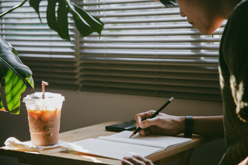 woman with short hair sits by the window, deeply engrossed in writing notes using pencil, on a book and iced coffee, in serene and focused atmosphere at home