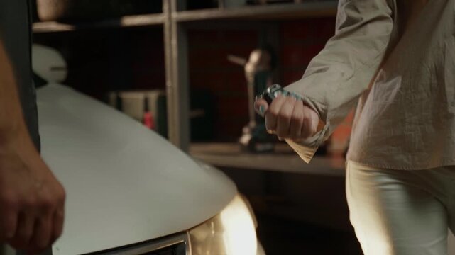 The mechanic hands over the car keys to the customer after the repair is completed in the garage. The mechanic is wearing a gray jumpsuit, and the client is wearing a beige shirt 