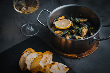 Mussels in a saucepan cooked in white wine with parsley and lemon on the table with a toasted baguette and a glass of white wine on a gray background