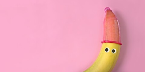 Banana with  googly eyes and red condom hat on pink background. Safe sex concept.	