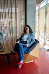 A 57-year-old woman sits with a laptop in a chair in a modern workspace