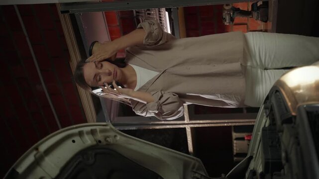 Vertical video: A woman in a brown shirt and white trousers is working on a car in her garage, holding a coffee cup and talking on her mobile phone