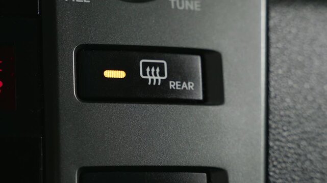 A close-up view of a rear window defroster button inside a car. The button is black and has a white icon of a window with three arrows pointing upwards. The word REAR is printed below the icon