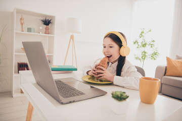 Photo portrait of cute small schoolgirl earphones online lesson eat lunch croissant netbook home education concept school interior
