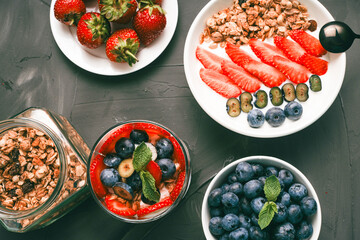 Berry parfait with granola and Greek yogurt in a white bowl and transparent glass on a gray textured background with strawberries and blueberries. Selected focus