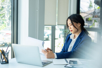 Businesswoman sitting at desk on couch in workplace or at home working on laptop and analyzing data on charts and graphs and writing on papers to make business plan 