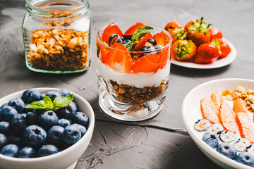 Dessert of strawberries and blueberries, granola and yogurt, cottage cheese and cream. A delicate parfait for a healthy breakfast in a white bowl and a glass glass with mint. Selected focus.