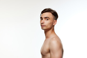 Shirtless young man with neatly styled hair looks confidently at camera, standing in profile...